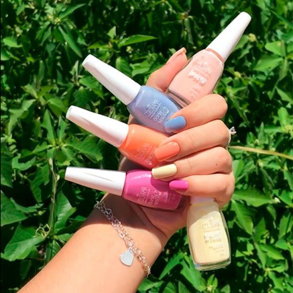 Mão segurando esmaltes coloridos em um fundo de folhagem verde.