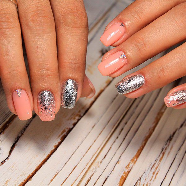 Unhas pintadas em tons de rosa e prata com detalhes de brilho sobre uma superfície de madeira clara.