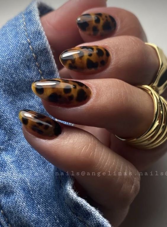 Unhas decoradas com estampa animal, segurando tecido jeans, com anéis dourados nos dedos.