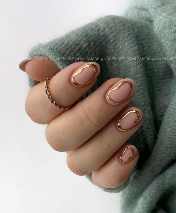 Mão com unhas decoradas em esmalte nude e bordas douradas, segurando um tecido cinza, anel dourado no dedo anelar.