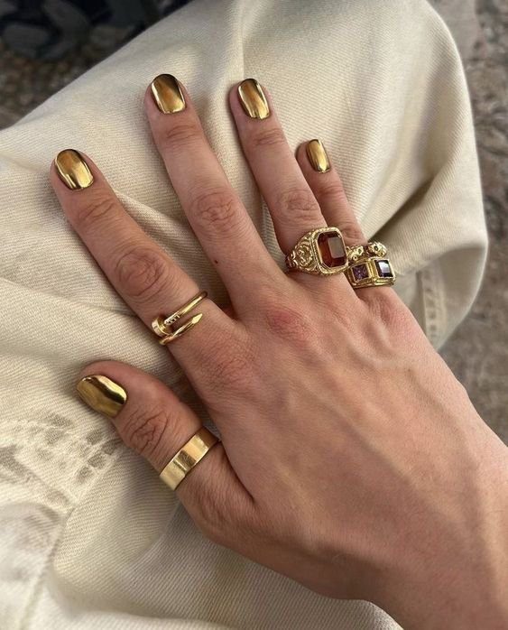 dourado-metalizado Mão com unhas douradas, usando anéis de ouro com pedras, repousando sobre tecido claro.