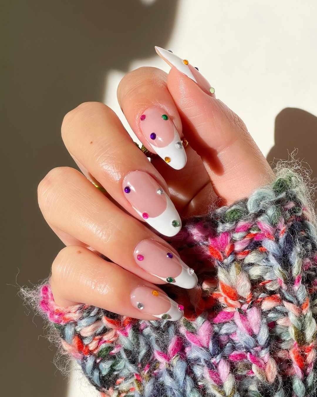 Mão com unhas decoradas em estilo francesinha, com esmalte branco e pedrinhas coloridas, segurando um tecido colorido.
