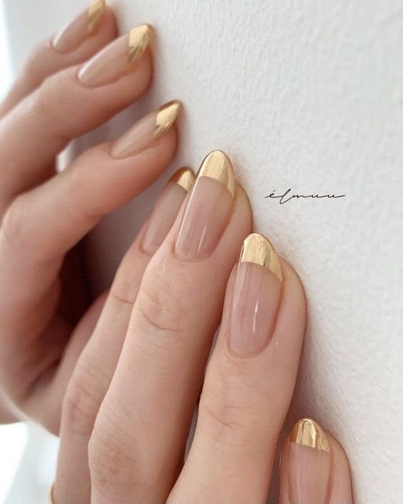french-manicure-dourada Mãos com unhas decoradas em nude e pontas douradas, próximas a uma superfície clara.