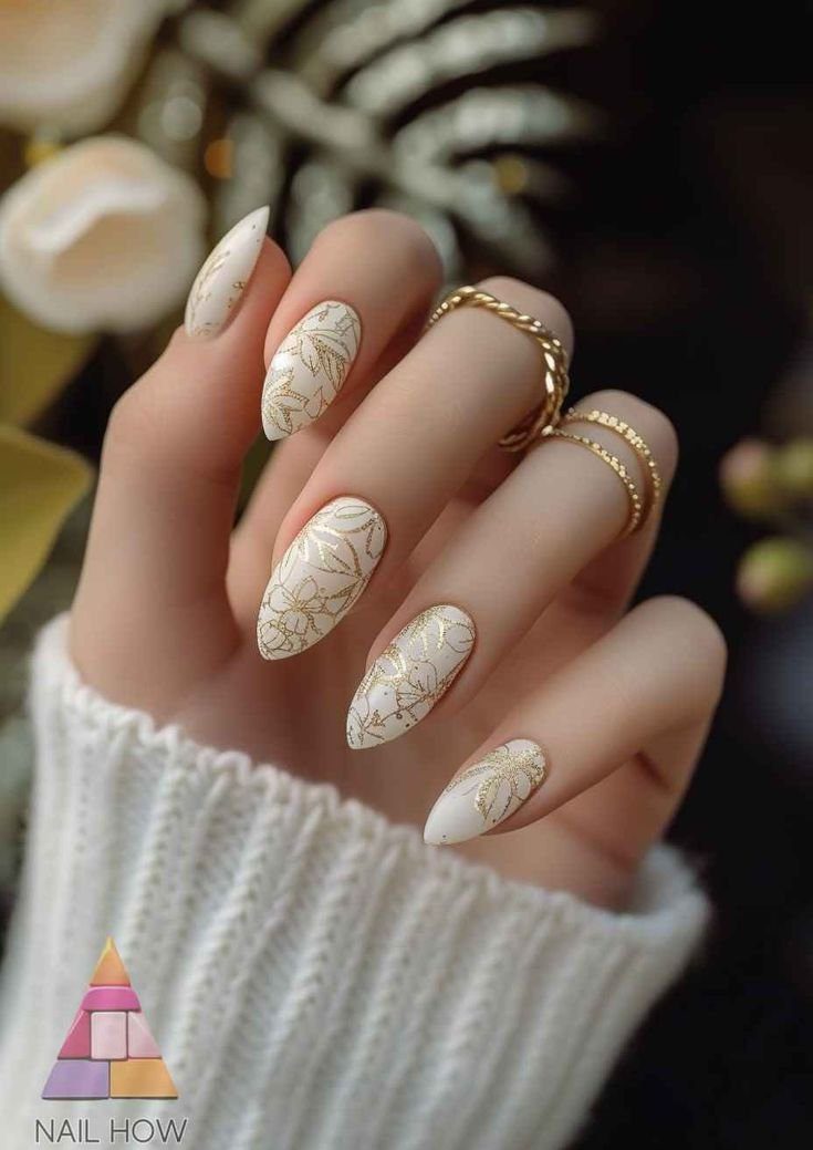 nail-stamping Mão com unhas decoradas em tons de branco e dourado, usando anéis finos dourados e vestindo um suéter claro.