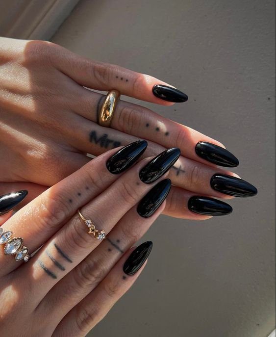 Duas mãos com unhas pretas longas e tatuagens nos dedos, usando anéis dourados e prateados.
