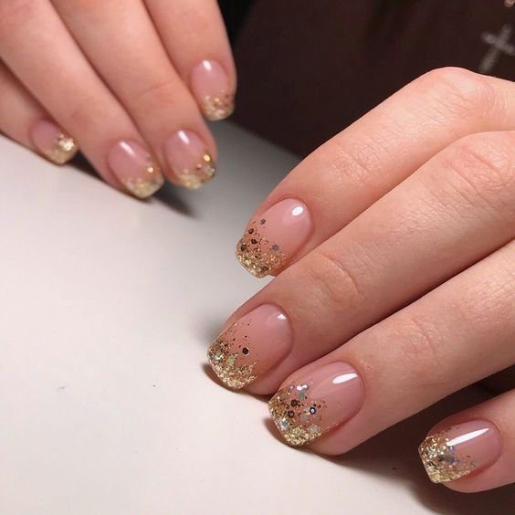 unha-decorada-com-glitter-dourado Mãos com unhas decoradas em esmalte nude e pontas com glitter dourado, sobre superfície clara.