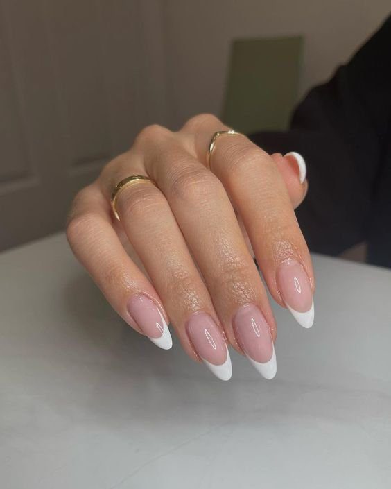 Mão com unhas decoradas em estilo francesinha, esmalte rosa claro e pontas brancas, usando anéis dourados.