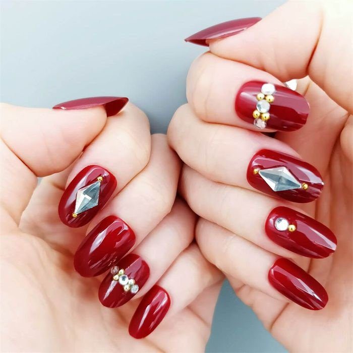 Mãos exibindo unhas pintadas de vermelho com decoração de pedras e detalhes brilhantes.