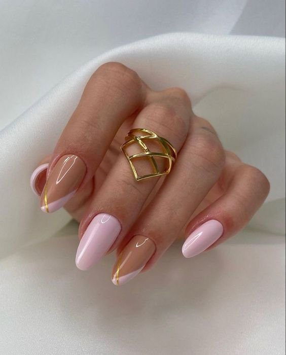 Mão com unhas longas e decoradas em tons de rosa e dourado, usando um anel dourado sobre tecido branco.