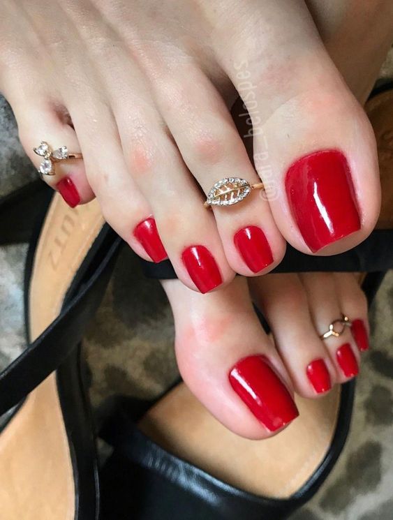 Pés com unhas pintadas de vermelho e adornados com anéis nos dedos, usando sandálias pretas.