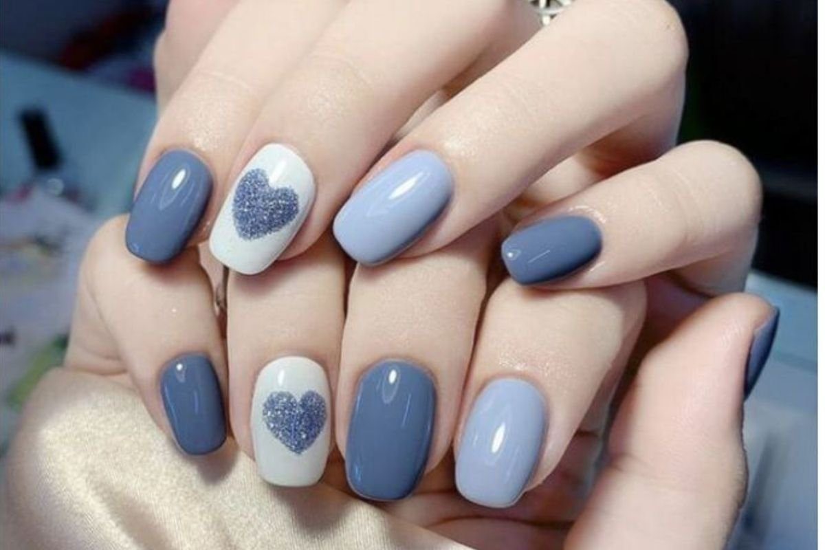 Unhas pintadas em tons de azul e branco, com detalhes de corações brilhantes em algumas unhas.