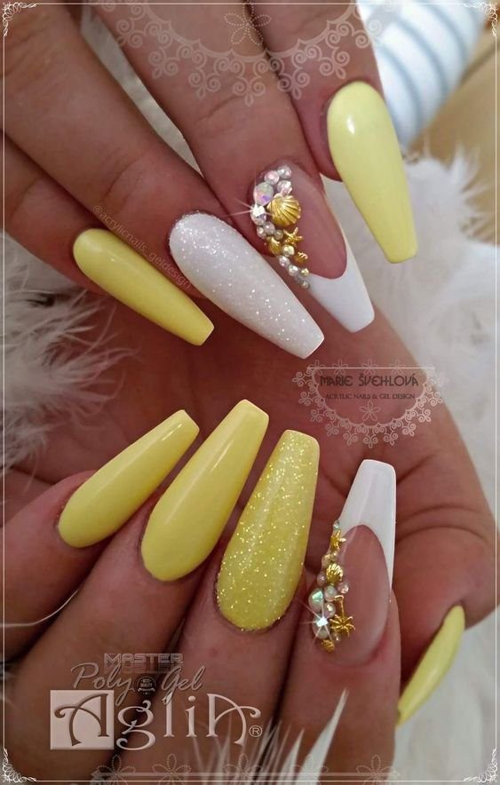 Unhas com esmalte amarelo e branco, decoradas com glitter e pequenas pedras brilhantes.