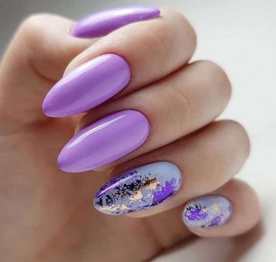 Unhas pintadas em tons de lilás e azul, com detalhes decorativos dourados e roxos.