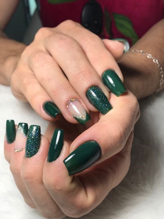 Mãos com unhas decoradas em esmalte verde escuro com detalhes brilhantes e uma unha com design geométrico.