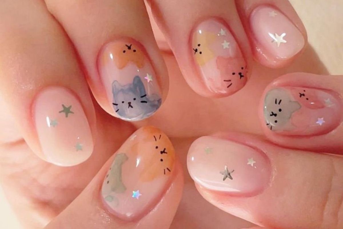 Unhas decoradas com desenhos de gatos coloridos e pequenas estrelas sobre fundo claro.