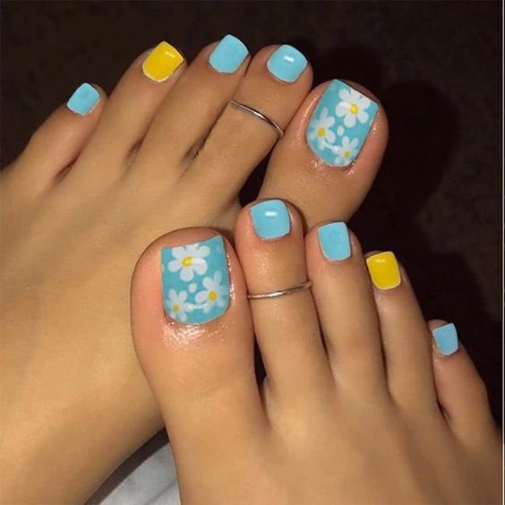 Unhas dos pés pintadas de azul e amarelo, com decoração floral, e anéis nos dedos.