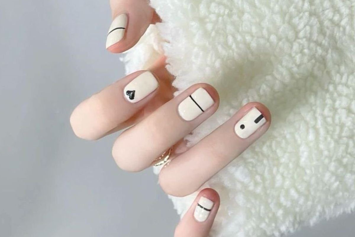 Mão com unhas decoradas em tons claros, com linhas pretas e um pequeno coração, sobre tecido felpudo branco.