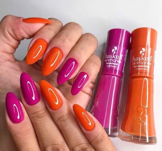 524d24e9dca229bdb8e1769c583e1e4e Mãos com unhas longas pintadas de rosa e laranja ao lado de dois frascos de esmalte nas mesmas cores.