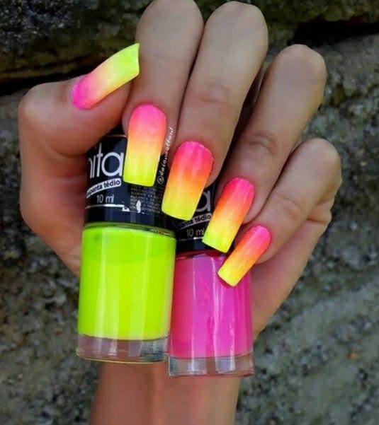 662fdf57dcce0b1812ae48b7500f8e22 Unhas compridas com esmalte em degradê de amarelo a rosa, segurando vidros de esmalte neon verde e rosa.