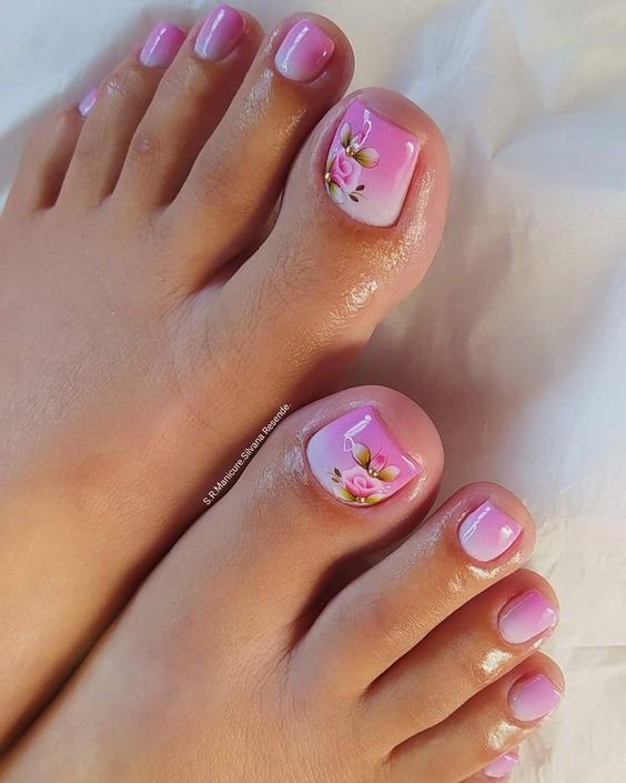 Pés com unhas pintadas de rosa claro, decoradas com flores na unha do dedão, sobre superfície branca.