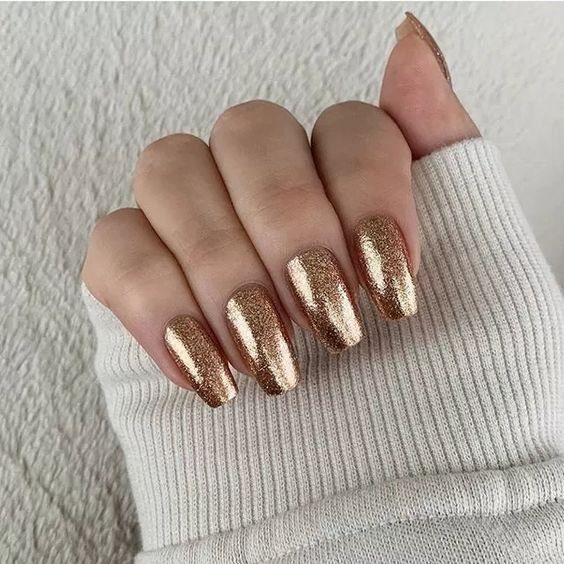 Mão com unhas longas e esmaltadas em tom metálico dourado, sobre tecido claro.