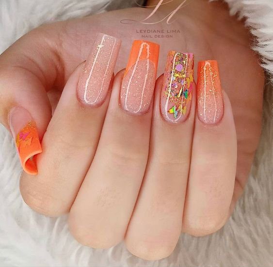 924b5cdf389f94bca3c52e72f35f2b65 Unhas decoradas com esmalte laranja, glitter e detalhes coloridos sobre fundo neutro.