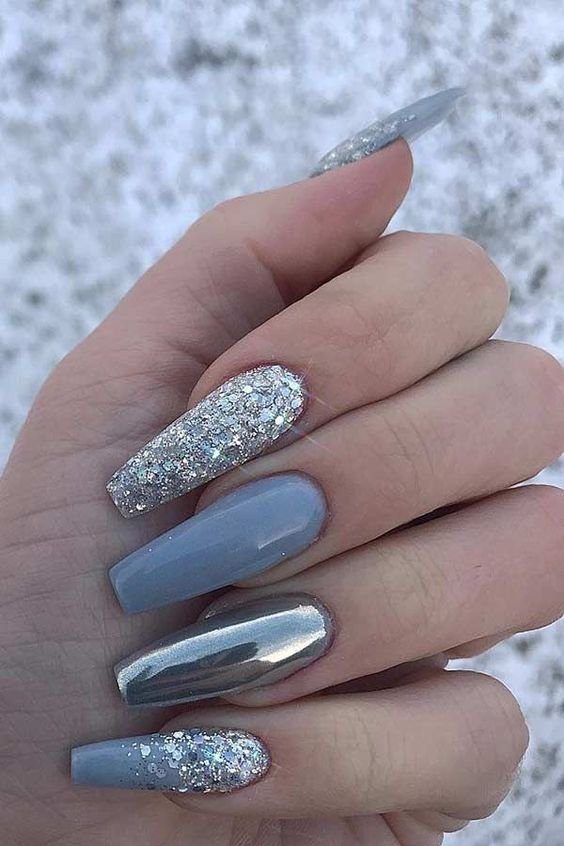 Mão com unhas longas e decoradas em tons de azul e prateado, com glitter e acabamento metálico.