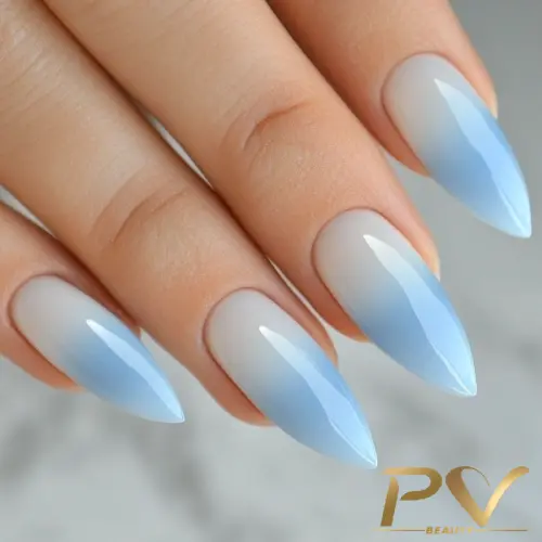 Unhas longas e pontiagudas com esmalte em degradê azul claro, sobre fundo neutro.