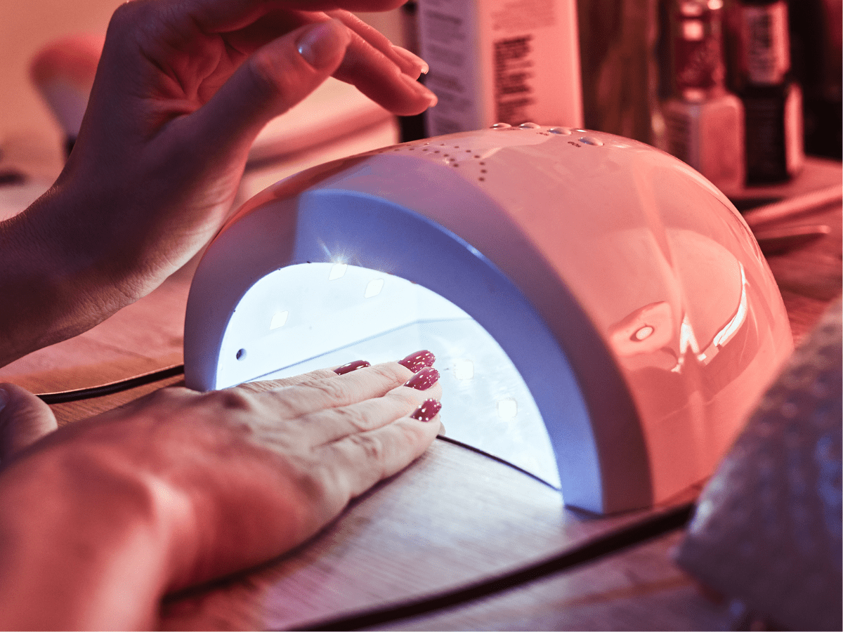 Mão com esmalte vermelho sendo seca em uma lâmpada LED para unhas sobre uma mesa de madeira.