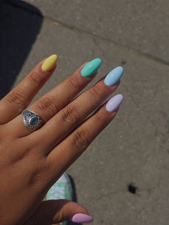 bbdff12e4b7941c71825a684d2bf03dc Mão com unhas coloridas em tons pastel e anel prateado, sobre fundo de concreto.