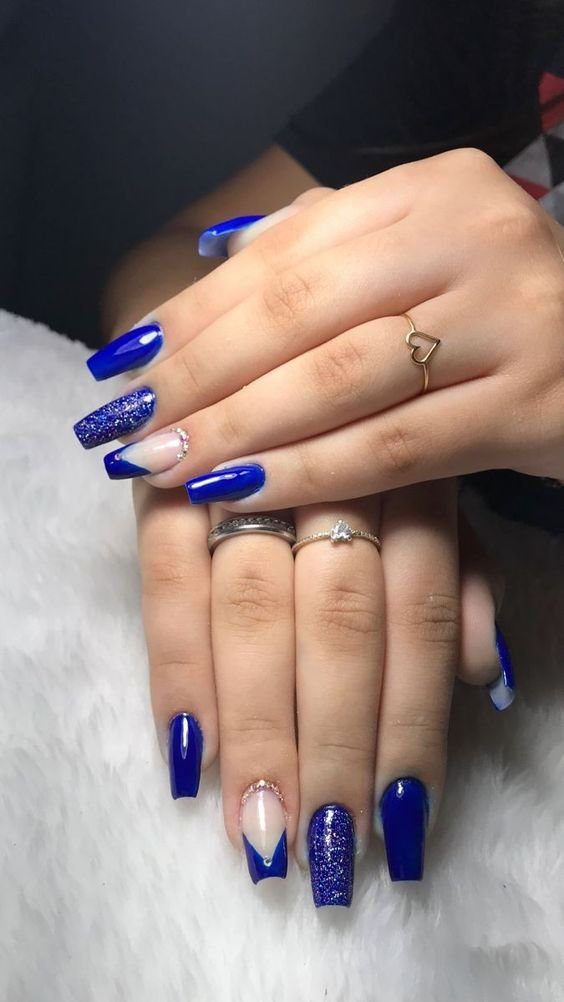 Mãos com unhas longas e decoradas em azul brilhante e detalhes prateados, com anéis delicados nos dedos.