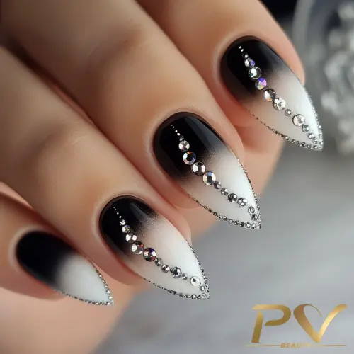 Unhas decoradas em preto e branco, com formato pontiagudo, adornadas com pequenas pedras brilhantes.