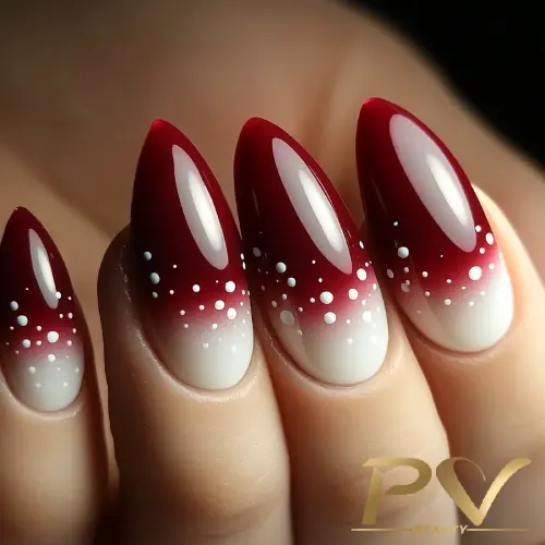 Unhas longas e pontiagudas com esmalte vermelho degradê e pequenas bolinhas brancas na superfície.