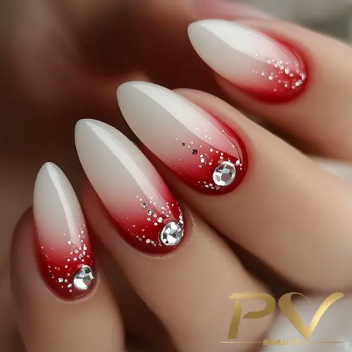 Unhas com esmalte degradê vermelho e branco, decoradas com pedras brilhantes e pequenos detalhes prateados.