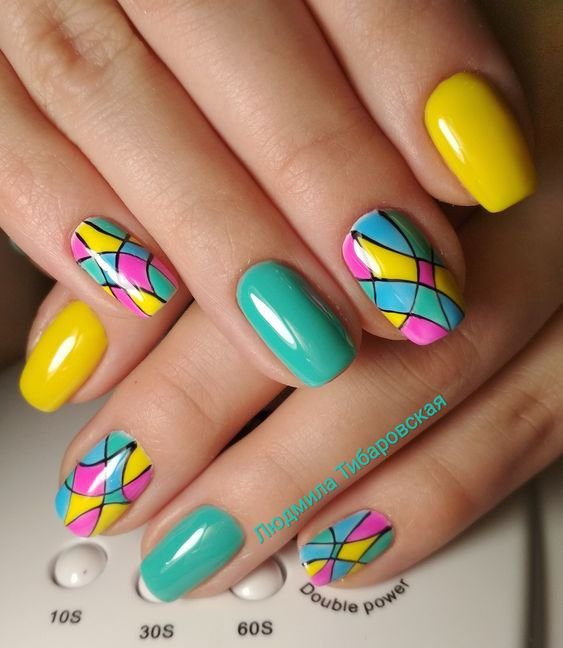detalhes-geometricos-nas-unhas Unhas decoradas com esmalte amarelo, azul e desenho colorido em padrão geométrico.