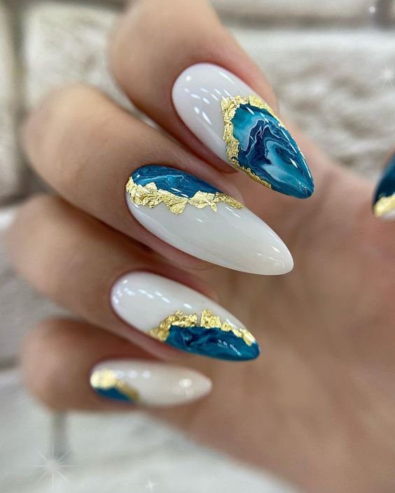 Unhas decoradas com esmalte branco, azul marmorizado e detalhes dourados.