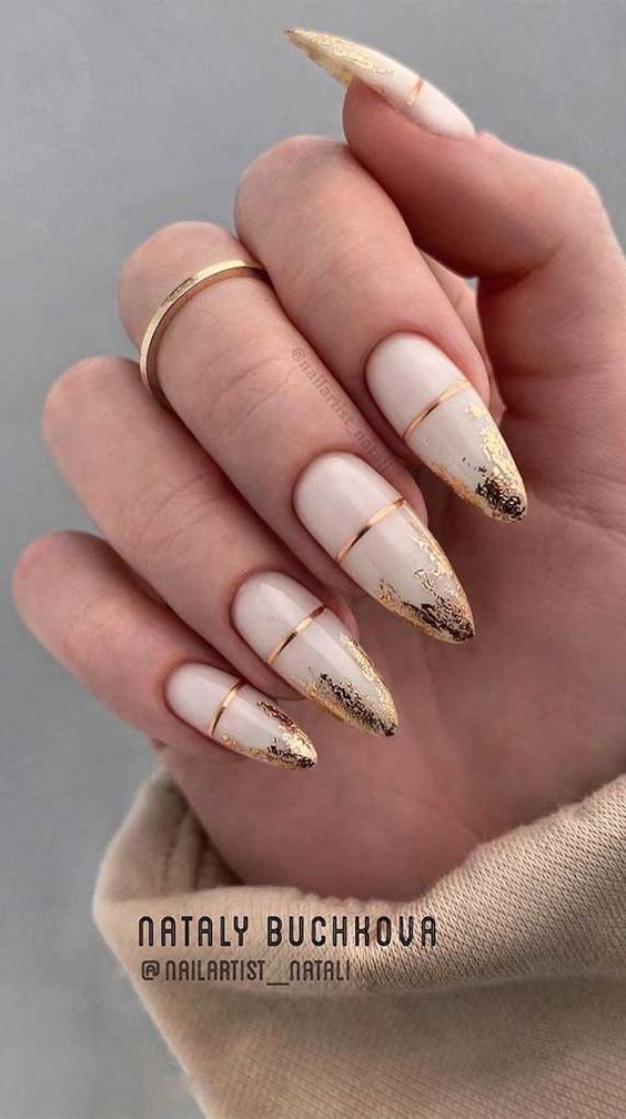 Mão com unhas compridas, decoradas com esmalte nude e detalhes dourados, destacando anéis finos nos dedos.