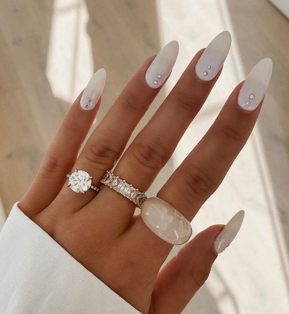 Mão com unhas longas brancas decoradas e três anéis brilhantes em ambiente iluminado.