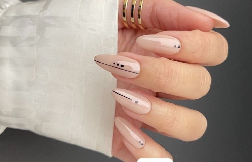 Mão com unhas alongadas, esmalte nude e decoração minimalista com linhas pretas e pequenos pontos.