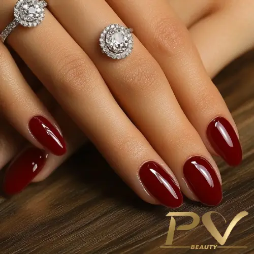 Mão com unhas longas pintadas de vermelho escuro e anéis prateados com pedras brilhantes sobre superfície de madeira.
