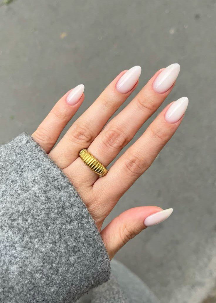 Mão com unhas pintadas de nude, usando anel dourado e vestindo casaco cinza.