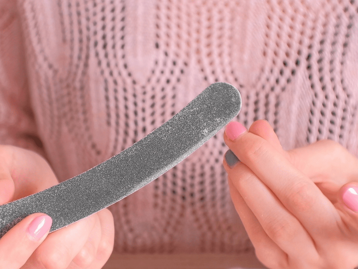 Mãos segurando uma lixa de unha cinza, com unhas pintadas de rosa e cinza, sobre um fundo de malha rosa.