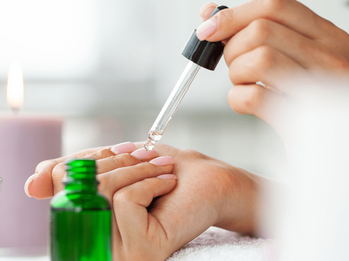 Mãos aplicando óleo em unhas com conta-gotas, ao lado de frasco verde e vela acesa.