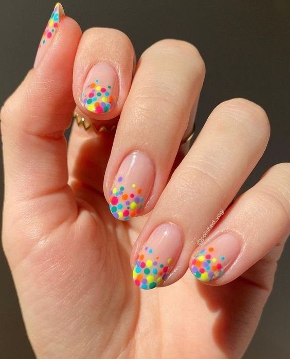 unhas-com-bolinhas-coloridas-para-o-carnaval Mão com unhas decoradas com esmalte transparente e pontinhos coloridos nas pontas em tons de azul, amarelo e vermelho.