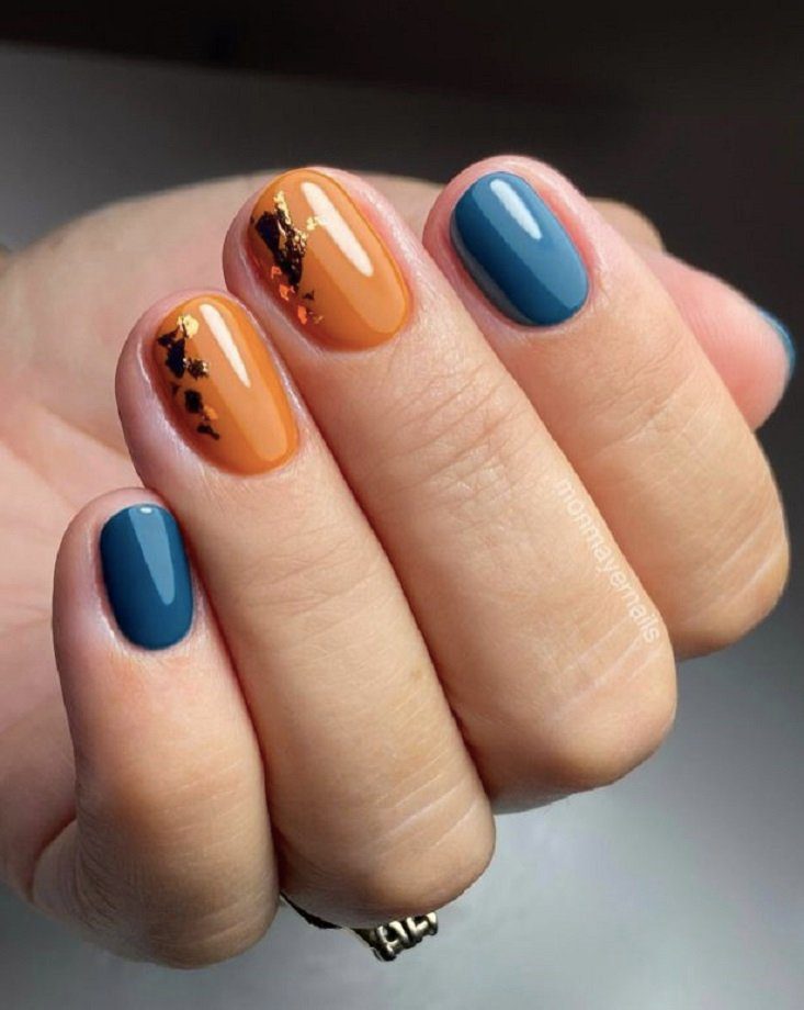unhas-curtas-coloridas-para-o-outono Mão com unhas pintadas em azul e laranja, duas com detalhes decorativos pretos e dourados.