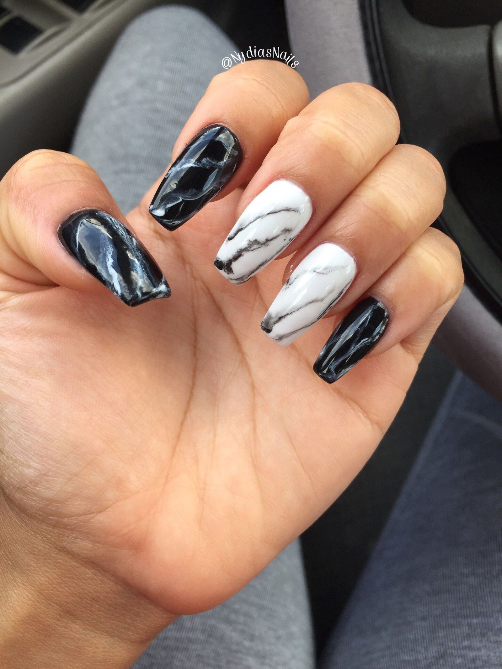 Unhas decoradas em preto e branco, com efeito marmorizado, são exibidas em uma mão segurando o volante de um carro.
