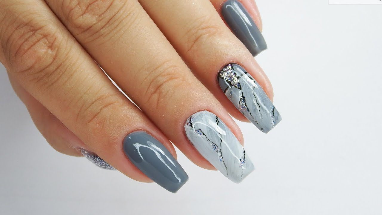 Mão com unhas decoradas em tons de cinza, com detalhes marmorizados e glitter.