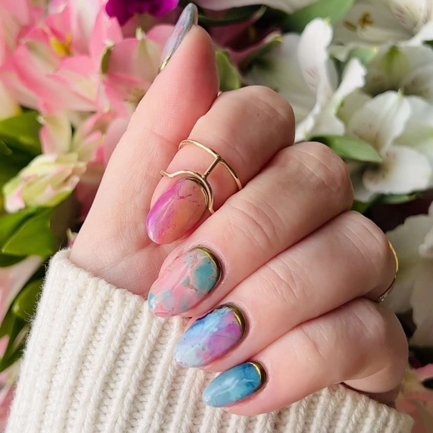 Mão com unhas decoradas em tons de rosa e azul, usando anéis, com flores ao fundo.