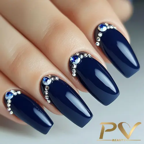 unhas-decorada-azul-strass-chique-elegante-pv-beauty-10 Unhas pintadas de azul escuro com pedras brilhantes decorativas na base, em uma mão com unhas longas e bem cuidadas.