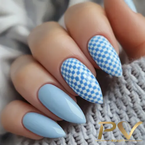 unhas-decorada-azul-xadrez-claro-pv-beauty-24 Mão com unhas longas e pontiagudas, esmalte azul claro e padrão xadrez azul e branco, sobre tecido cinza.